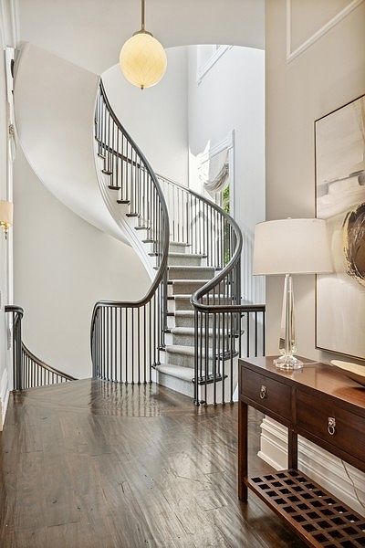 This elegant foyer features a dramatic curved staircase with dark railings and light-colored carpeted steps, serving as the central architectural focal point. To the right, a wooden console table topped with a glass-based lamp and a large abstract artwork creates a sophisticated entry experience. The space is finished with dark wood flooring and neutral walls, illuminated by a warm, spherical pendant light hanging from the upper level.