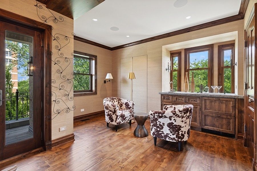 This elegant sitting area or study features rich wood flooring and textured wall coverings, creating a sophisticated atmosphere. Two cow-print armchairs are positioned around a small metallic side table, complemented by a built-in wet bar with dark wood cabinetry and a stone countertop. Large windows provide ample natural light and views of the outdoors, while a glass-paned door leads to an adjacent balcony.