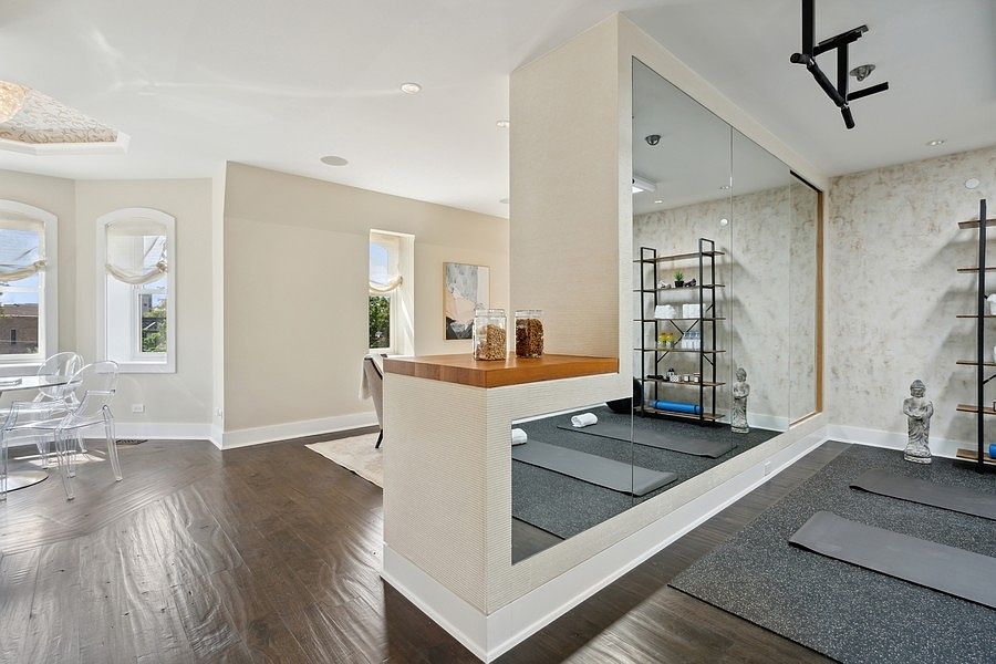 This multi-functional space features a dedicated home gym area with a large wall mirror, yoga mats, and open shelving for equipment. Adjacent to the gym, a dining area with a glass table and transparent chairs creates a seamless transition between fitness and living spaces. The room is characterized by dark wood flooring, neutral wall tones, and large windows that provide ample natural light.