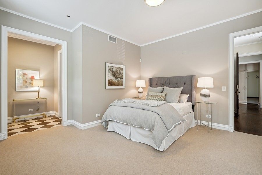 This elegant primary bedroom features a neutral color palette with a tufted gray headboard as the focal point. The room is symmetrically arranged with matching bedside tables and lamps, flanked by doorways leading to adjacent spaces, including a hallway with a distinctive black-and-white checkered floor. The space is well-lit with soft ambient lighting, creating a calm and sophisticated atmosphere.