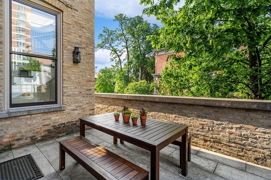 This outdoor terrace features a dark wood dining table and matching benches set against a rustic brick wall. The space is accented with small potted plants and overlooks a lush, tree-filled neighborhood, creating a private and serene atmosphere. The perspective is a medium shot from the terrace, capturing both the architectural details of the building and the surrounding greenery.