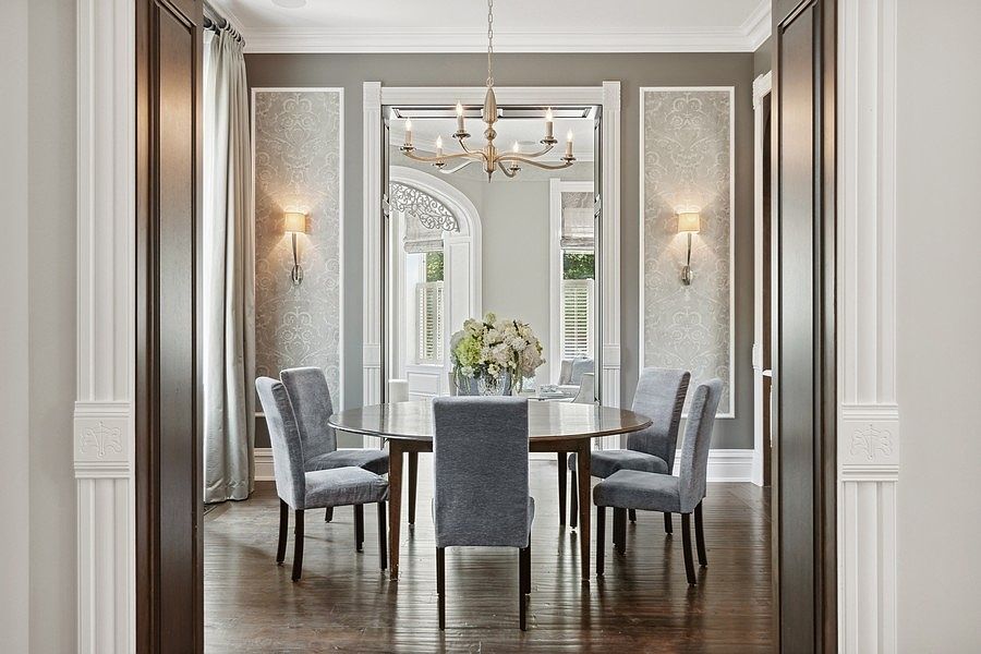 This elegant dining room is framed by dark wood-trimmed doorways, creating a sophisticated transition into the space. The room features a round wooden table surrounded by six upholstered chairs, centered beneath a classic chandelier. Decorative wall panels with patterned wallpaper and wall-mounted sconces add a refined touch, while a large mirror reflects the bright, airy windows beyond.