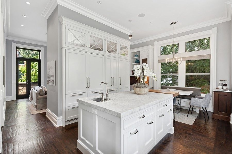 This bright, elegant kitchen features crisp white cabinetry with glass-fronted upper cabinets and a central island topped with a light-toned stone surface. A cozy breakfast nook with a built-in bench sits beside large windows that provide ample natural light and a view of the outdoors. The space is finished with dark hardwood flooring, creating a sophisticated contrast against the light cabinetry and walls.
