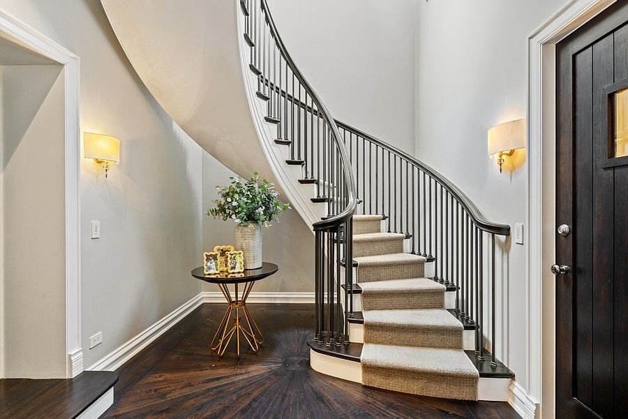 This elegant entryway features a dramatic curved staircase with a dark wood handrail and a neutral carpet runner. A round pedestal table with a floral arrangement and framed photos sits on the dark wood flooring, which displays a decorative circular inlay pattern. The space is illuminated by two wall-mounted sconces, creating a warm and inviting atmosphere that leads toward a dark-toned front door.