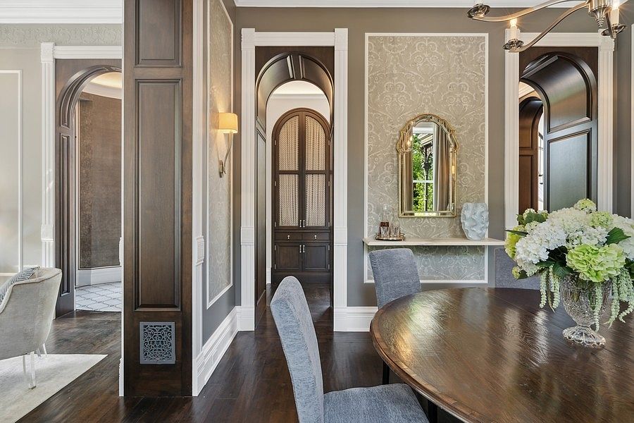 This elegant dining room features a dark wood circular table surrounded by upholstered chairs, set against a backdrop of sophisticated architectural details. The space is defined by arched doorways with dark wood trim, patterned wallpaper panels, and a decorative mirror above a floating shelf. The perspective captures the refined transition between rooms, highlighting the rich contrast between the dark wood flooring and the neutral wall tones.