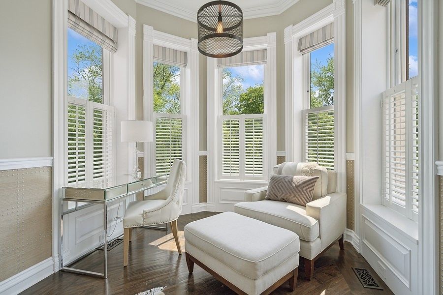 This bright and elegant home office or reading nook features a bay window arrangement that floods the space with natural light. The room is furnished with a mirrored desk, a tufted chair, and a comfortable armchair with a matching ottoman, all set against a backdrop of textured wall paneling and dark wood flooring. A modern, dark-toned drum pendant light hangs centrally, providing a sophisticated contrast to the light-colored furniture and window shutters.