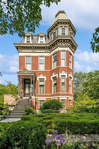 This striking Victorian-style brick residence features a prominent corner tower with a rounded roof and intricate architectural detailing. A grand stone staircase leads to an ornate, covered entryway, while the front yard is meticulously landscaped with manicured boxwood hedges and purple allium flowers. The perspective is a low-angle, eye-level shot that emphasizes the home's verticality and historic charm.