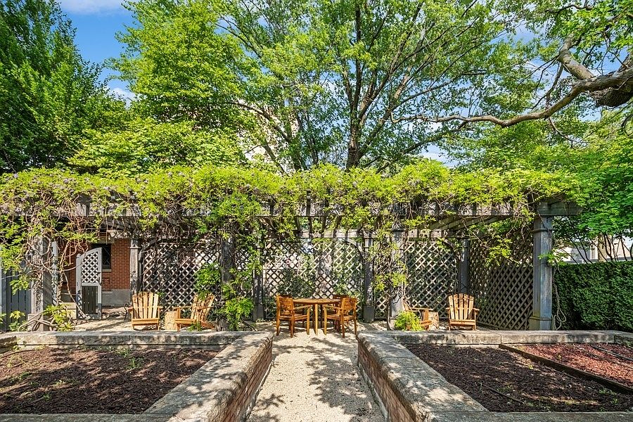 This serene garden space features a classic wooden pergola draped in lush greenery, providing a shaded seating area with a central table and chairs. The foreground includes symmetrical raised garden beds with stone borders, leading toward a gravel path that invites visitors into the tranquil outdoor retreat. The scene is framed by mature trees and lattice fencing, creating a private and peaceful atmosphere perfect for relaxation.