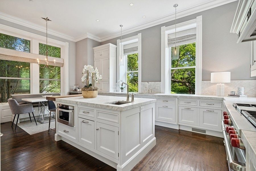 This bright and elegant kitchen features white cabinetry, a large central island with a stone countertop, and dark wood flooring. The space is illuminated by pendant lighting and natural light streaming through tall windows that overlook lush greenery. A cozy breakfast nook with a built-in bench is tucked into the corner, creating a warm and inviting atmosphere.