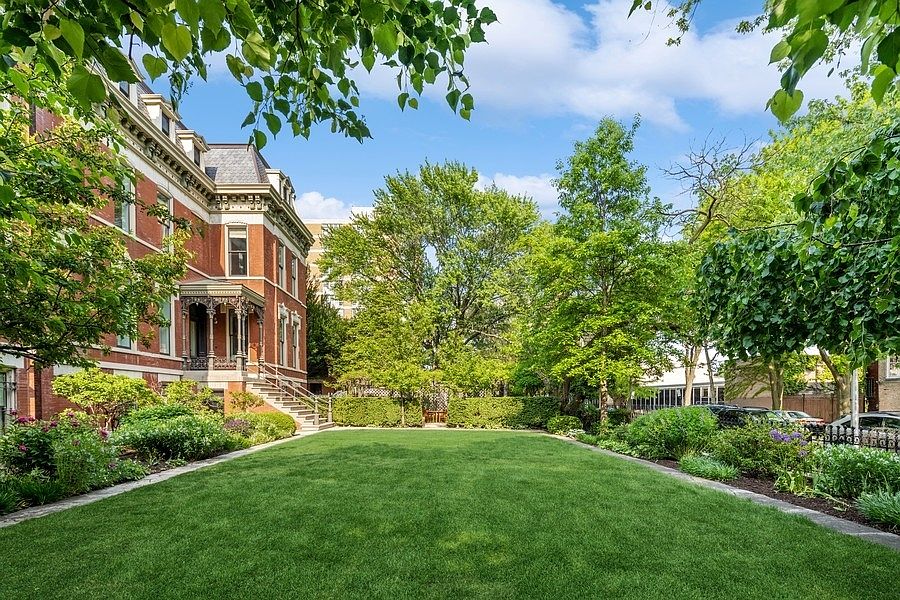 This image captures a lush, expansive green lawn framed by mature trees and manicured hedges, providing a serene outdoor retreat. To the left, a stately brick building with ornate architectural detailing and a covered porch adds a sense of historic elegance to the scene. The perspective is a wide-angle shot from the lawn looking toward the building, emphasizing the generous size of the private garden space.