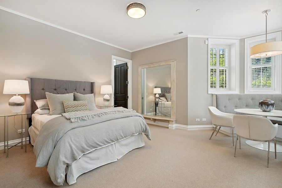 This spacious primary bedroom features a neutral color palette with a plush tufted headboard and a cozy seating area near the window. A large, framed floor mirror reflects the room, while a round dining table with two chairs creates a versatile nook for morning coffee or reading. The room is well-lit by a central ceiling fixture and matching bedside lamps, offering a serene and sophisticated atmosphere.