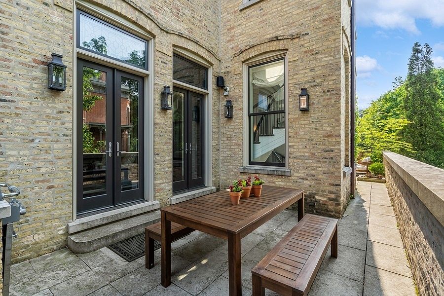 This outdoor patio space features a classic brick exterior wall with dark-framed French doors and a tall window. A wooden dining table with two matching benches sits on the stone-paved terrace, accented by small potted plants. The area is bordered by a stone retaining wall, offering a private and inviting setting for al fresco dining.
