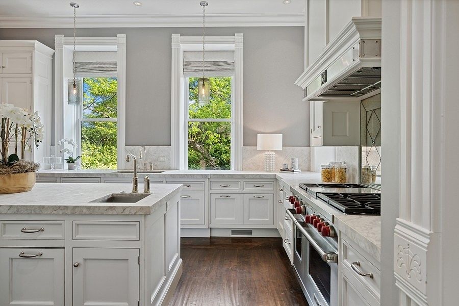 This elegant kitchen features crisp white cabinetry, a large central island with a stone countertop, and a professional-grade range with a prominent vent hood. Two tall windows allow natural light to flood the space, highlighting the dark wood flooring and the neutral gray wall color. The design is sophisticated and classic, with pendant lighting and a clean, organized aesthetic that emphasizes high-end finishes.