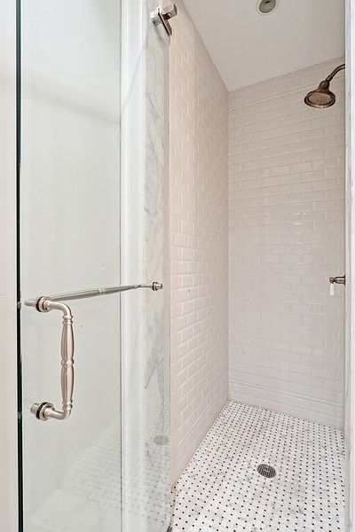 This shower enclosure features classic white subway tile walls and a decorative patterned tile floor with a central drain. A glass door with a polished metal handle provides entry, while a wall-mounted shower head is positioned above. The space is bright and clean, showcasing a timeless design aesthetic.