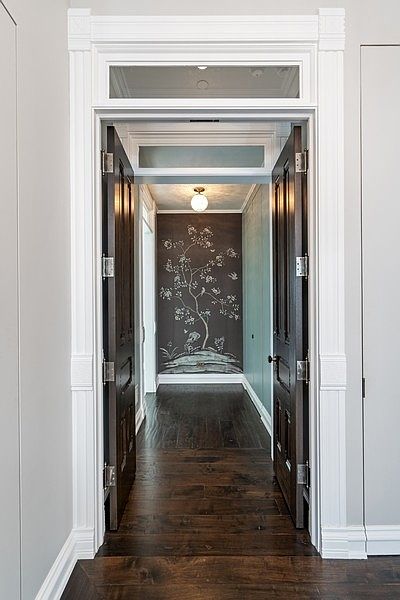 This image captures a view through a pair of dark, paneled double doors into a sophisticated hallway. The entryway features ornate white trim and a transom window above the doors, leading to a space accented by a dark wall mural depicting a stylized tree. A single globe pendant light illuminates the dark wood flooring, creating a dramatic and elegant transition between rooms.
