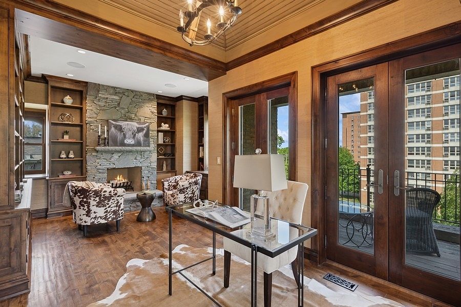 This sophisticated home office features a stone-clad chimney breast with a built-in fireplace and flanking wooden bookshelves. A glass-topped desk sits in the foreground, complemented by a tufted chair and a cowhide rug, while double glass doors open onto a balcony with an urban view. The room is finished with warm-toned wall coverings and a coffered wood ceiling, creating a refined and professional atmosphere.