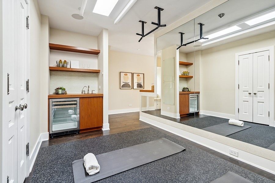 This home gym features a dedicated workout area with a rubberized floor mat and a wall-mounted pull-up bar. A built-in wet bar with a beverage refrigerator and open shelving provides convenient refreshment access, while a large wall mirror enhances the sense of space. The room is finished with neutral walls and dark wood flooring, creating a functional and polished environment for fitness.