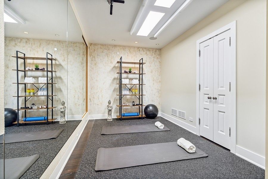 This dedicated home gym features a sleek, modern aesthetic with a large wall-to-wall mirror that enhances the sense of space. The room is equipped with two open-frame metal shelving units, two yoga mats laid out on the floor, and a black exercise ball, creating a functional and organized workout area. A white double door provides access to the room, while the neutral walls and textured flooring contribute to a calm, professional atmosphere.