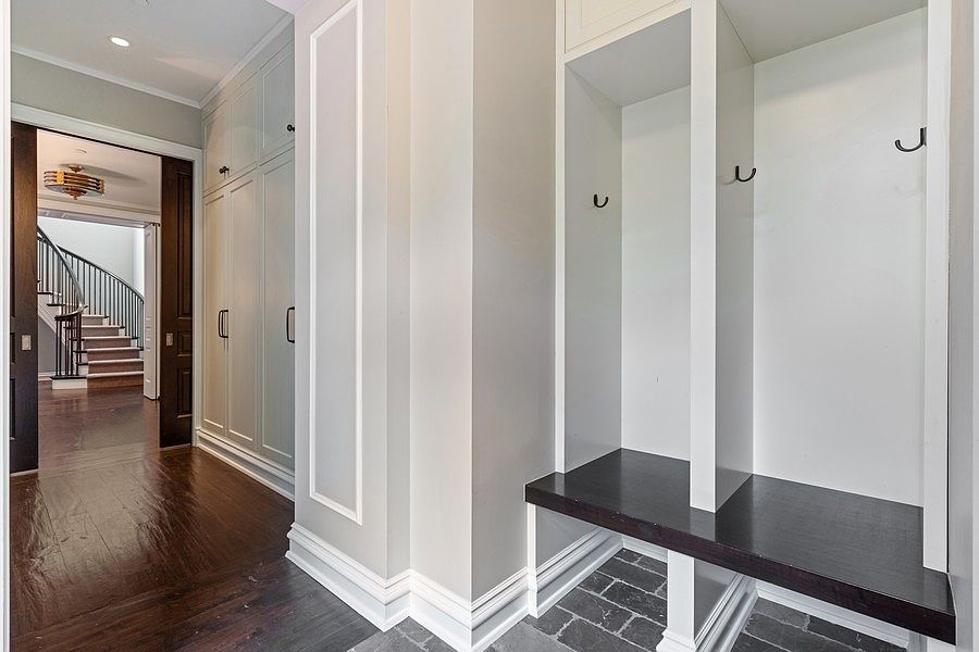 This interior view captures a mudroom area featuring built-in storage lockers with dark wood benches and black hooks, transitioning into a hallway with light-colored cabinetry. The flooring shifts from dark wood planks in the background to a dark stone tile in the foreground. Through the open doorway, a staircase with a curved railing is visible, creating a sense of depth and architectural flow.