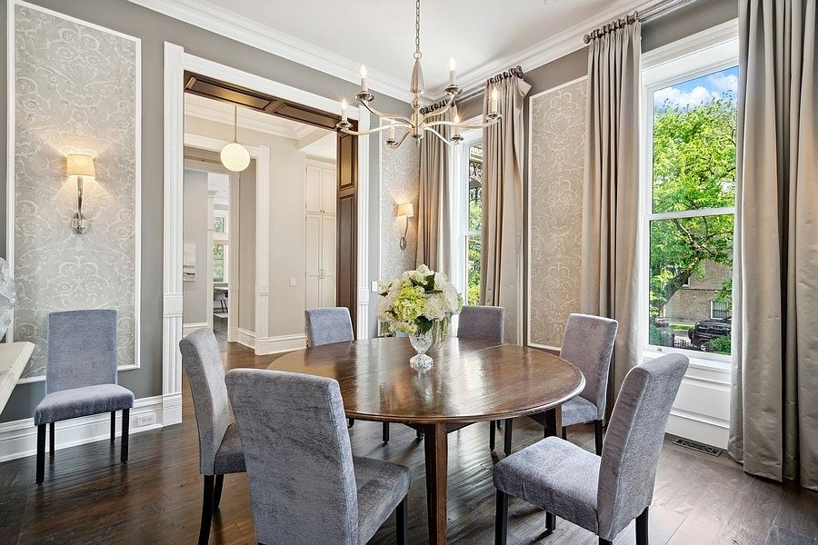 This elegant dining room features a round wooden table surrounded by six upholstered gray chairs, centered beneath a classic multi-armed chandelier. The space is defined by sophisticated wallpaper panels, tall windows draped in neutral curtains, and dark wood flooring. A wide doorway leads into an adjacent room, creating an open and airy flow throughout the home.