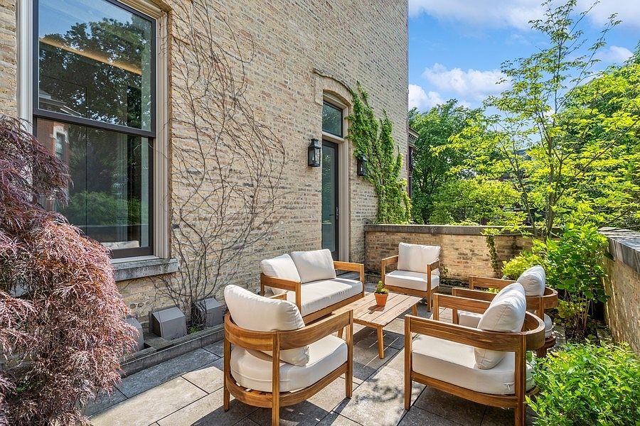 This inviting outdoor patio features a light-colored brick wall adorned with climbing vines, creating a charming and private atmosphere. The space is furnished with a modern wood-framed seating set with plush white cushions, centered around a low coffee table. Large windows and a glass-paneled door provide a seamless connection to the interior, while lush greenery and trees frame the stone-paved terrace.