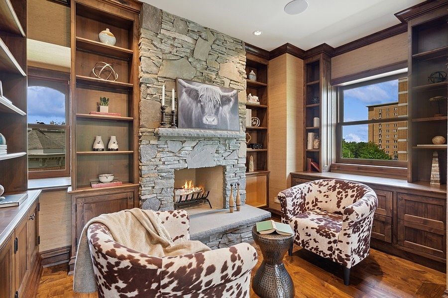 This cozy study features a prominent stone-clad chimney breast with a built-in fireplace and a decorative cow portrait above the mantel. The room is framed by extensive dark wood built-in shelving on either side, complemented by two cow-print armchairs and a small metal side table. Large windows on both sides of the fireplace provide natural light and views of the exterior, creating a warm and inviting atmosphere.