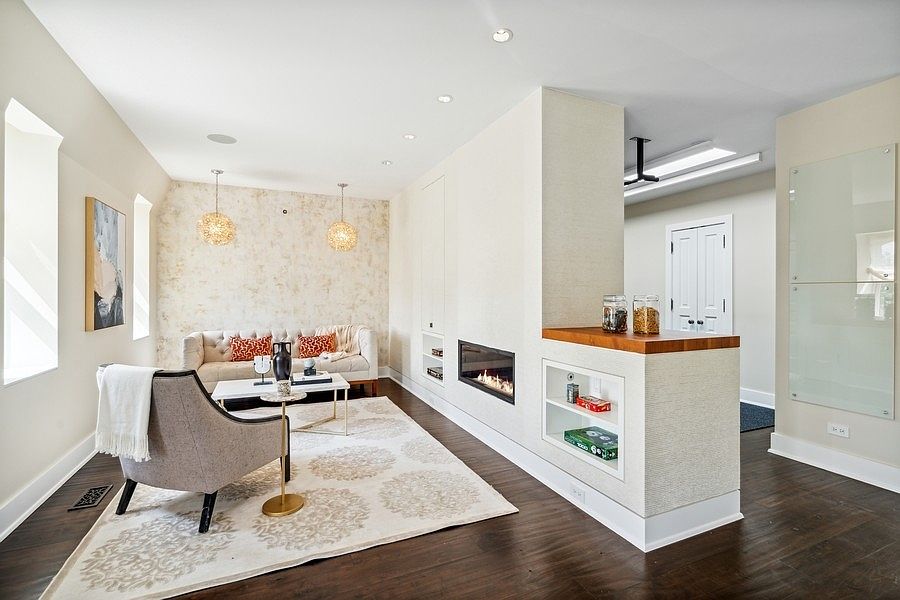 This inviting living room features a neutral color palette with a textured accent wall behind a tufted sofa and two decorative pendant lights. A modern linear fireplace is integrated into a central partition wall, which also includes built-in shelving and a wood-topped counter. The space is anchored by a large patterned area rug over dark wood flooring, creating a warm and sophisticated atmosphere.