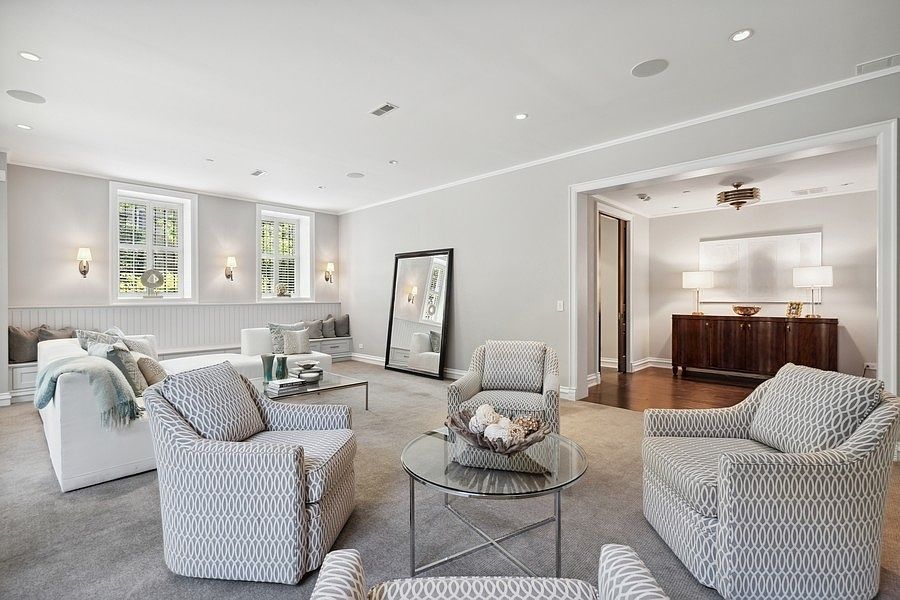 This spacious and elegant living room features a neutral color palette with plush carpeting and a sophisticated seating arrangement. The room is anchored by a large white sofa and several patterned armchairs, complemented by a glass-topped coffee table and a tall, leaning floor mirror. A wide opening leads to an adjacent room with a dark wood sideboard, creating an open and airy flow throughout the living space.