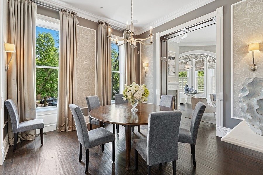 This elegant dining room features a round wooden table surrounded by six grey upholstered chairs, centered beneath a classic multi-armed chandelier. The space is defined by tall windows draped in neutral curtains, patterned wallpaper, and dark wood flooring that flows into an adjacent living area. The perspective captures the sophisticated transition between rooms, highlighting the refined architectural details and balanced lighting.