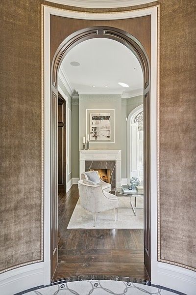 This perspective captures a view through a grand, arched doorway into a sophisticated living space. The foreground features walls covered in a textured, neutral-toned fabric, while the interior room showcases dark wood flooring, a fireplace with a stone surround, and elegant seating. The transition between the two spaces creates a sense of depth and architectural refinement, highlighting the home's high-end finishes.