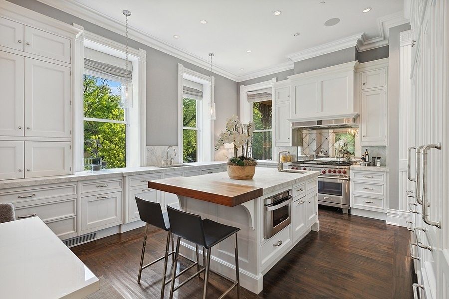This bright and elegant kitchen features white cabinetry, stone countertops, and a central island with a wood-topped breakfast bar and two stools. The space is illuminated by recessed lighting and two pendant lights hanging over the island, while large windows provide ample natural light and views of the outdoors. The dark wood flooring provides a rich contrast to the light-toned cabinetry and walls, creating a sophisticated and inviting atmosphere.