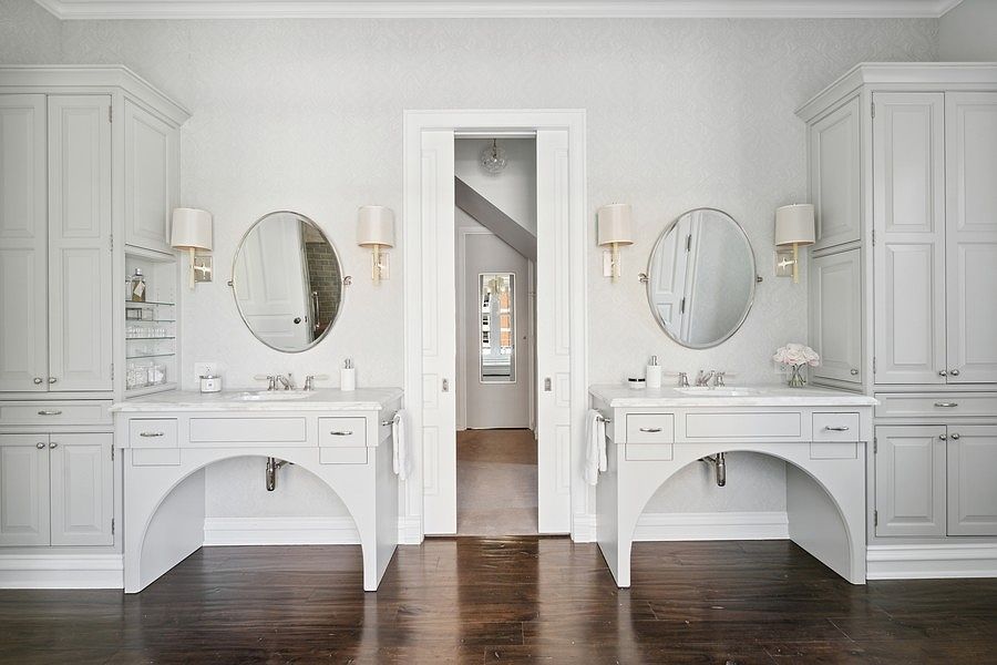 This symmetrical primary bathroom features dual vanity stations with light-toned cabinetry, stone countertops, and arched open bases. Each station is accented by a round mirror and wall-mounted sconces, set against a subtly patterned wallpaper. A central doorway leads to an adjacent hallway, creating a balanced and elegant layout with dark wood flooring throughout.
