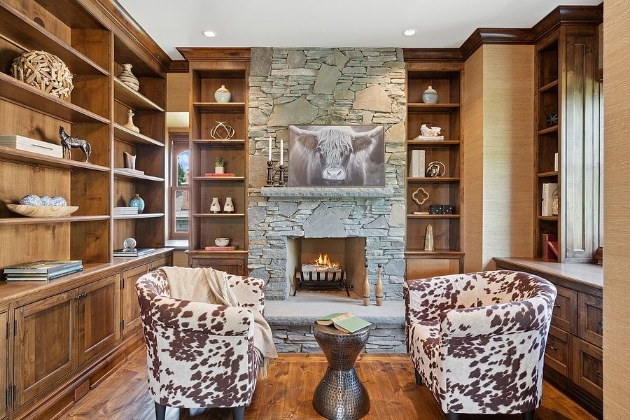 This cozy study features custom wood shelving units flanking a prominent stone-clad chimney breast that houses a warm fireplace. Two patterned armchairs face a metallic pedestal table, creating an inviting seating area centered in the room. The space is finished with rich wood flooring and warm, textured wall coverings, offering a sophisticated and rustic atmosphere.