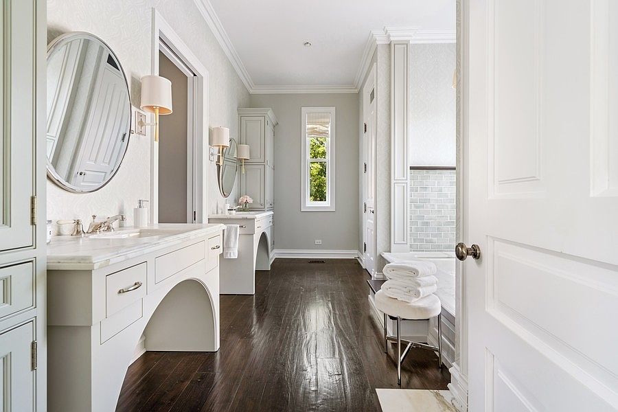 This elegant primary bathroom features a double vanity with light-toned cabinetry, stone countertops, and circular mirrors. The space is anchored by dark wood flooring that contrasts with the light walls, leading toward a window at the end of the room and a tiled bathtub area on the right. A small stool with folded towels sits near the tub, creating a serene and sophisticated atmosphere.