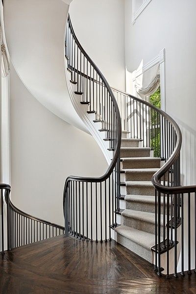 This elegant interior features a grand, curved staircase with dark wood handrails and black metal balusters. The steps are covered in a neutral-toned carpet, contrasting with the rich, dark wood flooring of the landing. The space is bathed in natural light from a tall window, creating a sophisticated and airy atmosphere.
