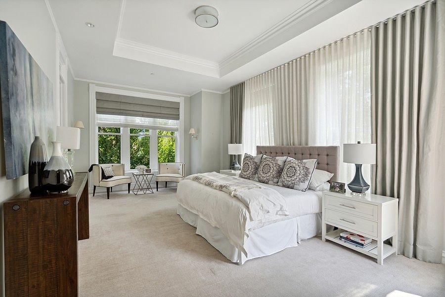 This spacious primary bedroom features a neutral color palette, a tray ceiling, and plush carpeting, creating a serene and sophisticated atmosphere. A large, upholstered bed serves as the focal point, flanked by white nightstands and matching lamps, while a separate seating area with two armchairs sits near the large windows. The room is bathed in natural light filtered through elegant, floor-to-ceiling drapery, offering a refined and comfortable retreat.