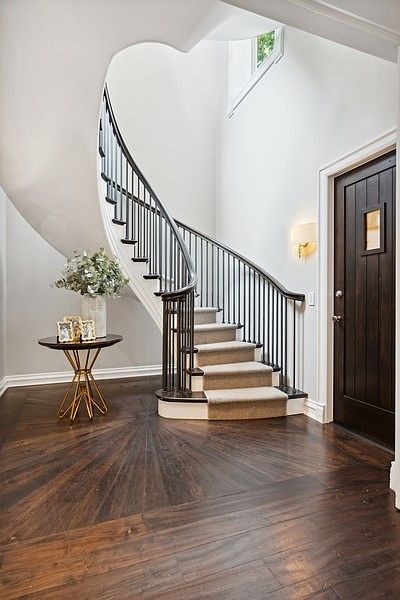 This elegant entryway features a dramatic curved staircase with a dark railing and a neutral runner, serving as the focal point of the space. The floor is finished in a striking radial wood pattern that draws the eye toward the stairs, complemented by a small, modern pedestal table with decorative accents. A dark wood door with a small window panel stands to the right, while a wall-mounted light fixture provides a warm glow against the crisp white walls.