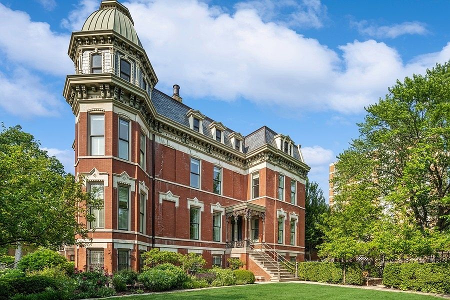 This image captures a grand, historic brick mansion featuring a prominent corner tower with a domed roof and intricate architectural detailing. The structure showcases a classic Second Empire style with a mansard roof, dormer windows, and ornate metalwork above the main entrance stairs. A well-manicured lawn and lush green landscaping frame the base of the building, creating an elegant and stately curb appeal.