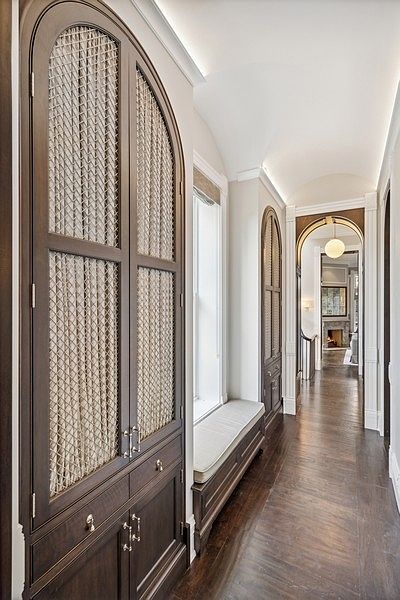 This elegant hallway features a long, arched built-in storage unit with decorative metal mesh door panels and integrated bench seating. The space is characterized by a barrel-vaulted ceiling, dark wood flooring, and a warm, neutral color palette that leads the eye toward a distant fireplace in the next room. The perspective is a low-angle, eye-level shot that emphasizes the depth and architectural symmetry of the corridor.