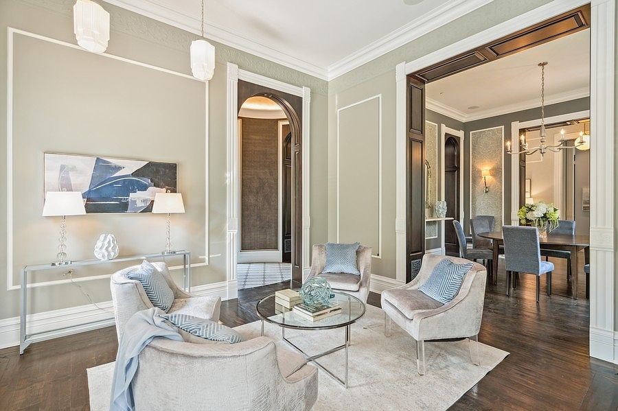 This elegant living room features a sophisticated neutral color palette with soft sage walls and dark wood flooring. The space is furnished with plush armchairs arranged around a circular glass coffee table, creating an inviting conversation area. A wide, dark-trimmed archway leads into an adjacent dining room, showcasing an open and airy floor plan with high ceilings and refined architectural molding.