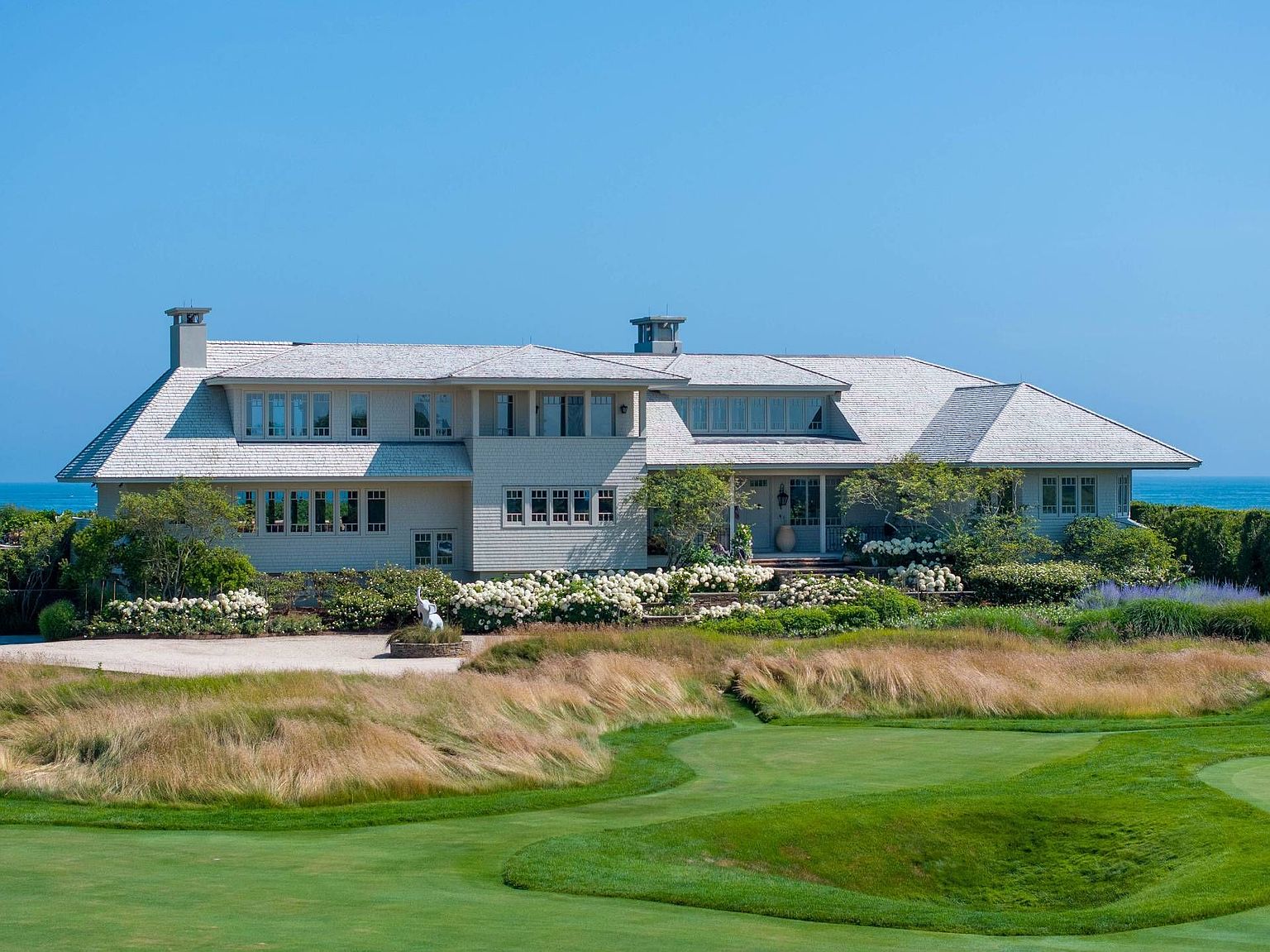 This expansive coastal estate features a classic shingle-style architectural design, characterized by its light-toned siding, multi-gabled roofline, and numerous windows that capture the surrounding landscape. The home is set against a backdrop of the ocean, with a meticulously landscaped garden filled with white hydrangeas bordering the foundation. In the foreground, a lush green golf course fairway transitions into tall, swaying grasses, creating a serene and picturesque setting for this luxury property.