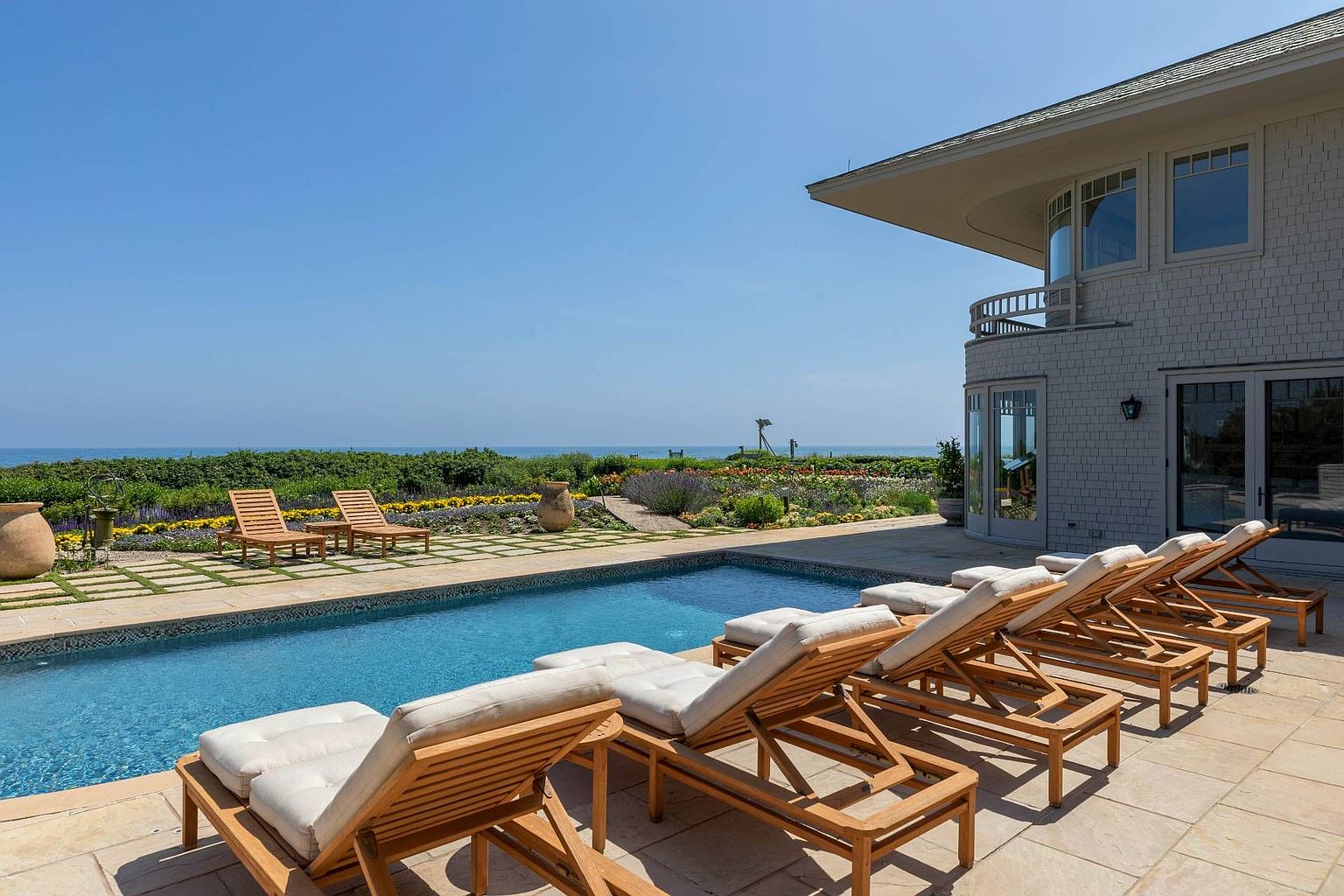 This expansive outdoor living space features a rectangular swimming pool lined with lounge chairs, set against a backdrop of a lush garden and the ocean horizon. The home showcases a shingled exterior with a curved architectural design and large glass doors that open onto a stone-paved patio. The scene captures a serene, high-end coastal lifestyle with a clear view of the landscape and the sea.
