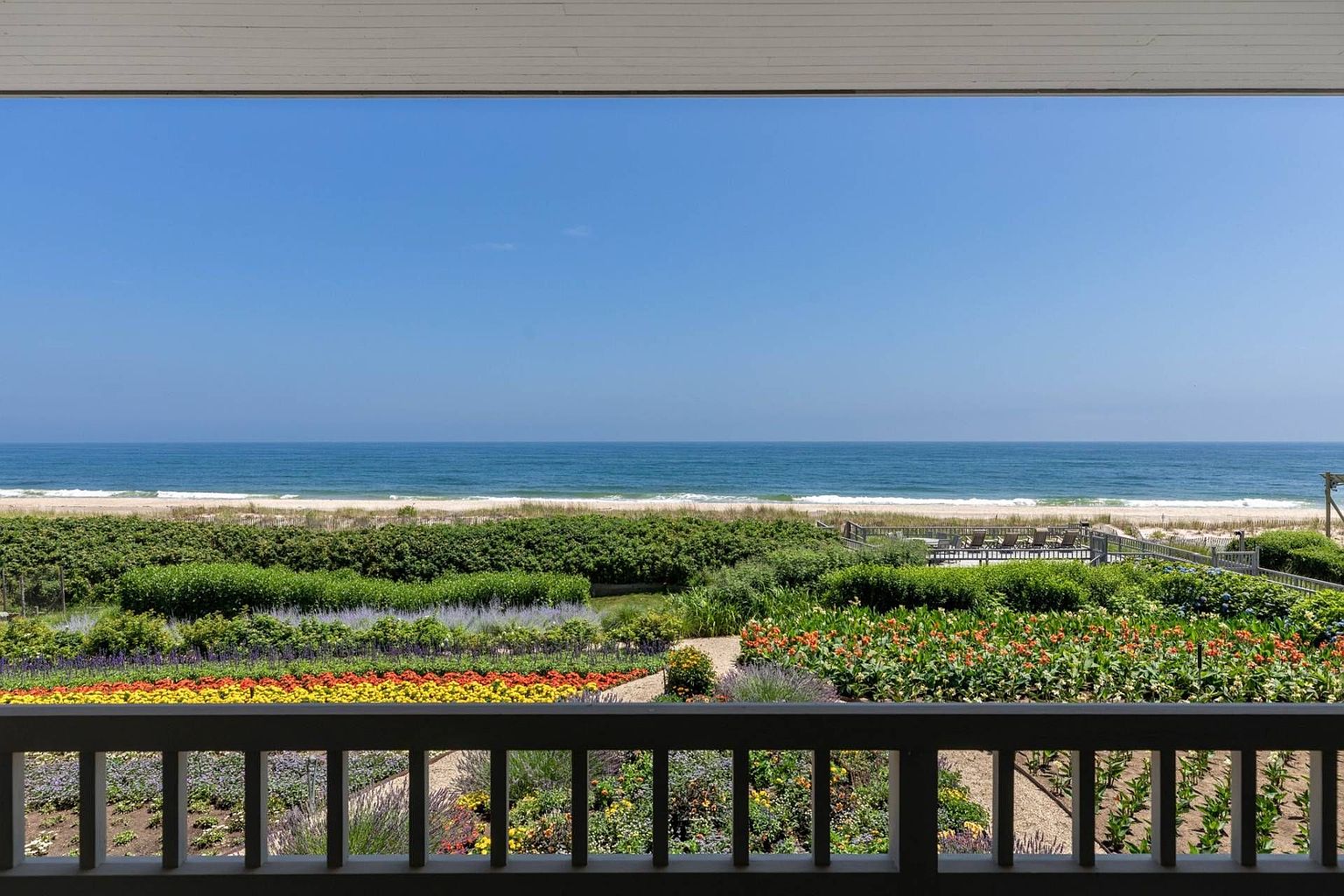 This view from a balcony captures a stunning, unobstructed panorama of the ocean and sandy beach. In the foreground, a vibrant, multi-colored flower garden adds a lush, manicured touch to the landscape, leading the eye toward the horizon. The perspective is elevated and serene, emphasizing the property's prime coastal location and seamless connection to nature.