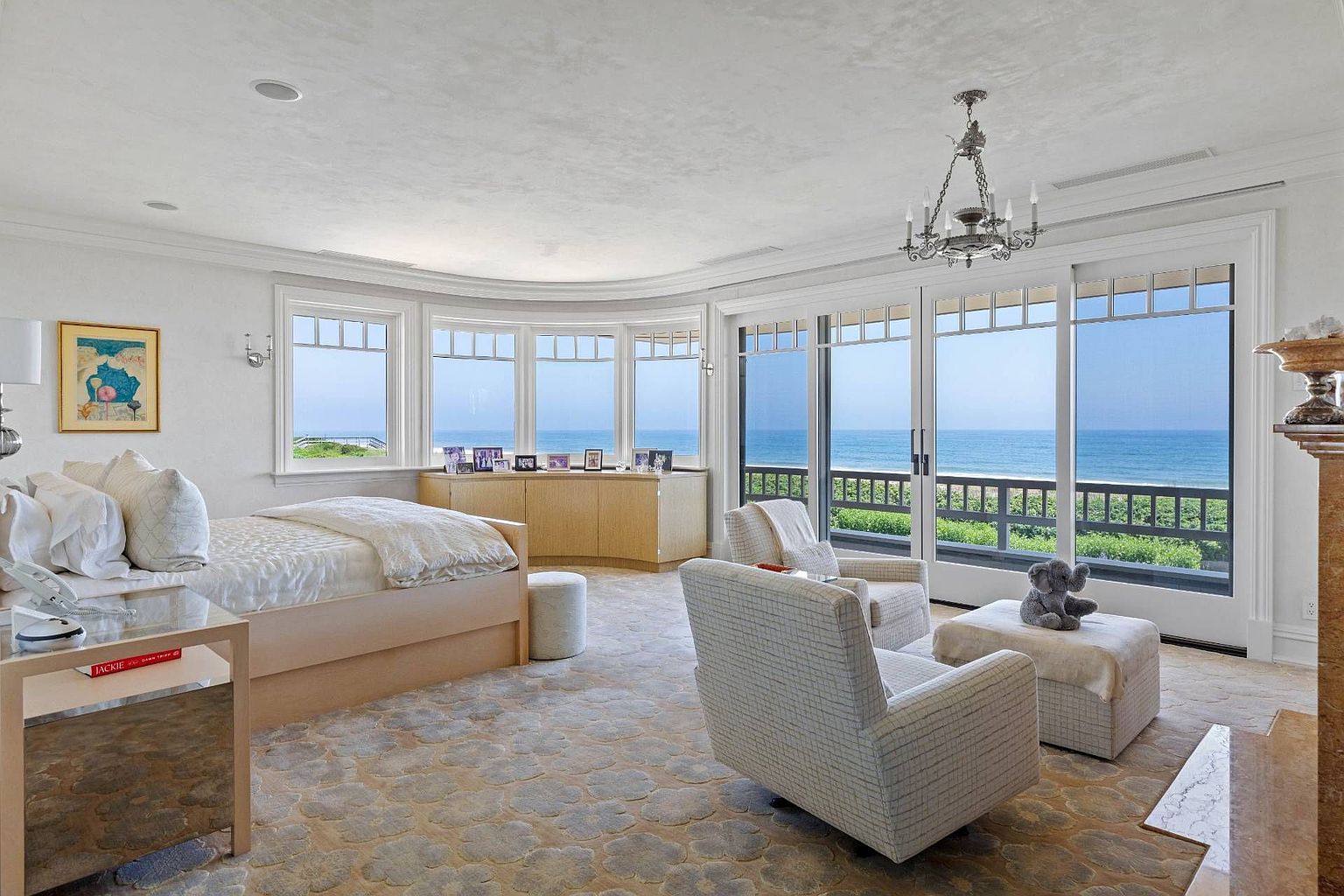 This spacious primary bedroom features a light-toned bed and a cozy seating area arranged near large glass doors that open to a scenic ocean view. The room is decorated in a soft, neutral palette with a patterned carpet and a classic chandelier, creating a serene and luxurious atmosphere. A curved built-in cabinet sits beneath the bay windows, providing storage while maintaining the room's elegant, airy aesthetic.