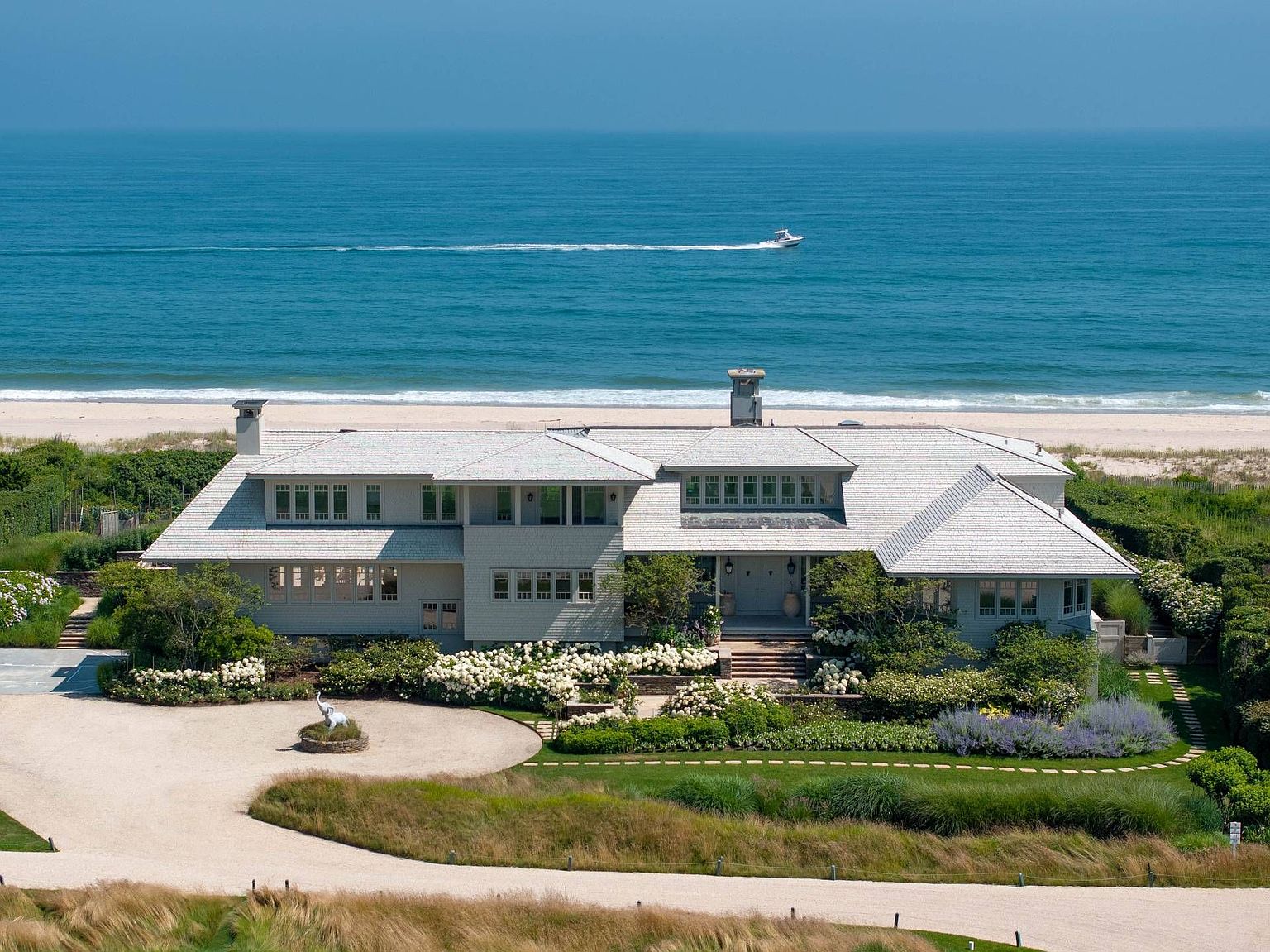 This stunning aerial perspective captures a sprawling, shingle-style coastal estate situated directly on the beachfront. The property features a light-colored exterior, a multi-gabled roofline, and lush, manicured landscaping with vibrant white hydrangea bushes framing the entrance. A circular gravel driveway leads to the home, while a small boat cruises across the calm, expansive blue ocean in the background.