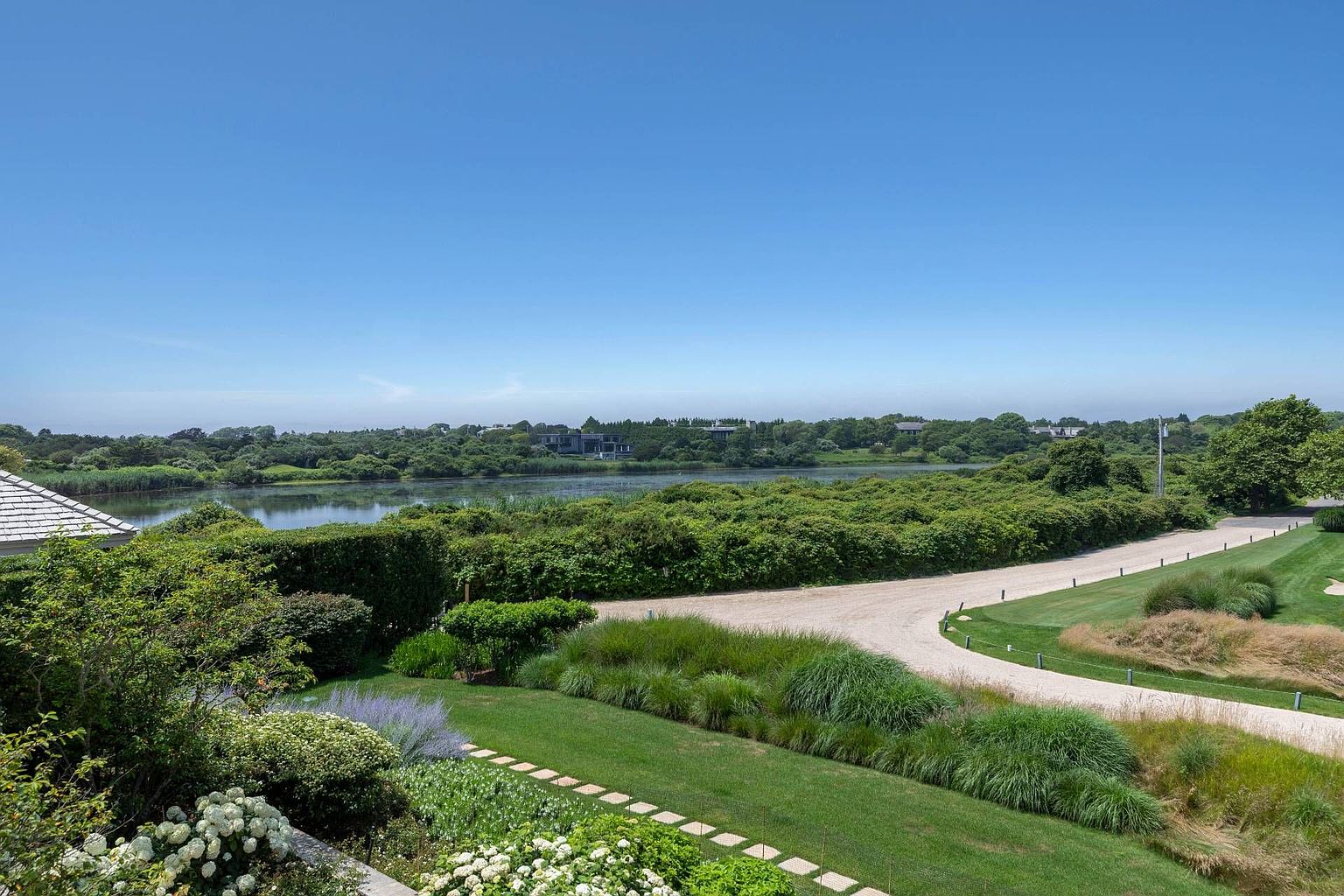 This elevated view captures a lush, expansive landscape featuring a manicured lawn, vibrant green shrubbery, and a winding gravel driveway. In the background, a serene pond reflects the clear blue sky, framed by dense trees and distant residential structures. The scene offers a peaceful, scenic perspective of the property's natural surroundings and outdoor space.