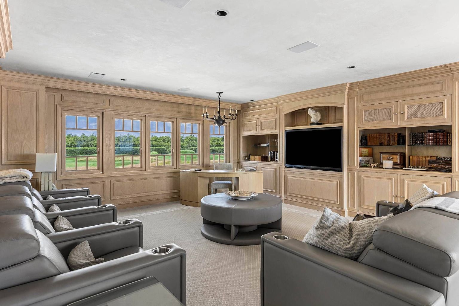 This sophisticated multi-purpose room features extensive light wood paneling and built-in cabinetry that houses a television and shelving. The space is furnished with a curved desk near a row of windows overlooking a green landscape, as well as a comfortable seating area with grey leather armchairs and a matching circular ottoman. The room offers a refined, professional atmosphere with a neutral color palette and a classic chandelier.