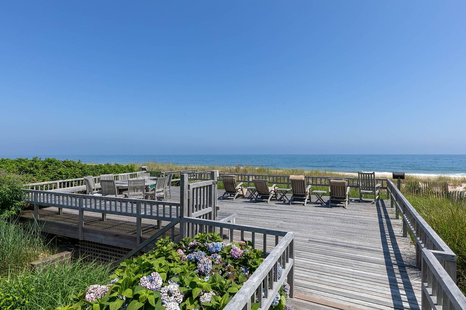 This expansive, weathered wood deck offers a prime vantage point overlooking the ocean and sandy dunes. The space is furnished with multiple outdoor dining and lounge chairs, creating an ideal setting for coastal entertaining. In the foreground, vibrant blue and purple hydrangea bushes add a touch of color to the natural, breezy aesthetic of this beachside retreat.