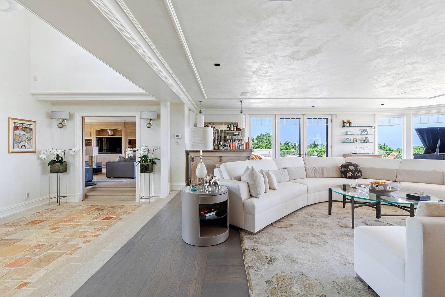 This spacious living room features a large, curved white sectional sofa arranged around a glass-topped coffee table on a patterned area rug. The room transitions from a stone-tiled entryway into a dark wood-floored living area, with a wet bar visible in the background and a glimpse into an adjacent den. Large glass doors provide ample natural light and a view of the outdoors, creating an open and airy atmosphere.