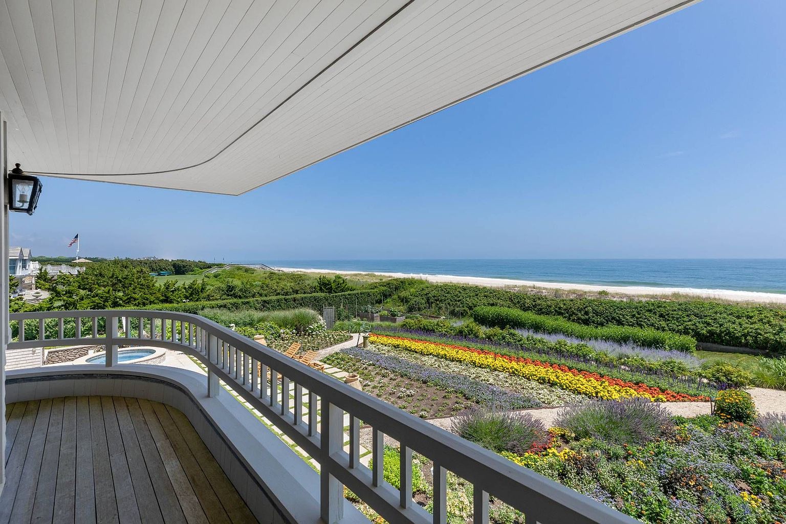 This elevated wooden balcony offers a sweeping, panoramic view of a vibrant, multi-colored flower garden that transitions into a lush green lawn and the sandy beach beyond. The scene is captured from a high vantage point, emphasizing the seamless connection between the private outdoor living space and the expansive coastal landscape. The bright, clear blue sky and the calm ocean horizon create a serene and luxurious atmosphere.